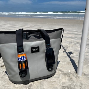 Puede incluir: Una bolsa de enfriador Yeti gris con correas negras y un soporte para tel&eacute;fono negro. La bolsa de enfriador est&aacute; sentada en una playa de arena con un oc&eacute;ano azul en el fondo. La bolsa de enfriador tiene un portavasos negro con una lata naranja dentro.