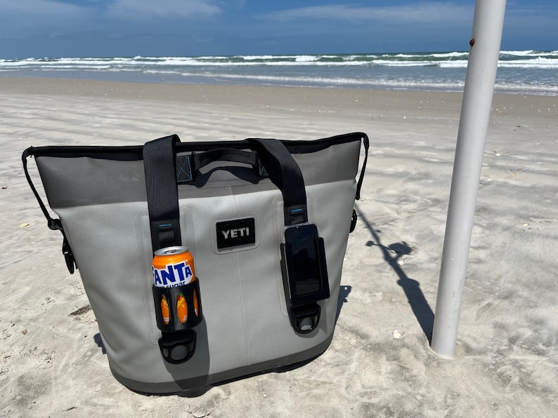 Puede incluir: Una bolsa de enfriador Yeti gris con correas negras y un soporte para tel&eacute;fono negro. La bolsa de enfriador est&aacute; sentada en una playa de arena con un oc&eacute;ano azul en el fondo. La bolsa de enfriador tiene un portavasos negro con una lata naranja dentro.