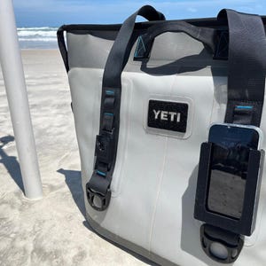 Puede incluir: Una bolsa de enfriador Yeti gris con un soporte de tel&eacute;fono negro adjunto al lado. El soporte de tel&eacute;fono tiene un tel&eacute;fono negro adentro. La bolsa de enfriador tiene una correa negra y un asa negra. La bolsa de enfriador est&aacute; sentada en una playa de arena blanca.
