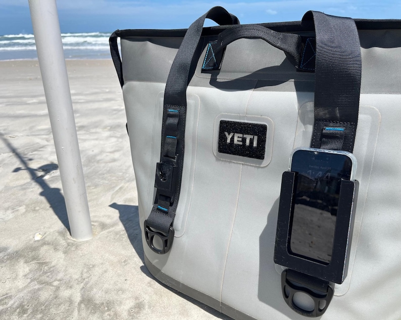 Puede incluir: Una bolsa de enfriador Yeti gris con un soporte de tel&eacute;fono negro adjunto al lado. El soporte de tel&eacute;fono tiene un tel&eacute;fono negro adentro. La bolsa de enfriador tiene una correa negra y un asa negra. La bolsa de enfriador est&aacute; sentada en una playa de arena blanca.