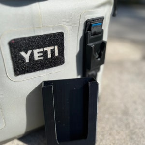 Puede incluir: Un soporte de tel&eacute;fono de pl&aacute;stico negro con una abertura en forma de U, fijado a una nevera blanca Yeti.