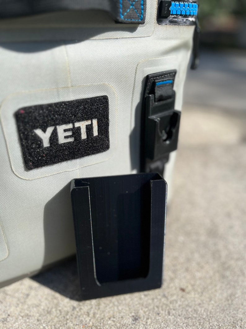 Puede incluir: Un soporte de tel&eacute;fono de pl&aacute;stico negro con una abertura en forma de U, fijado a una nevera blanca Yeti.