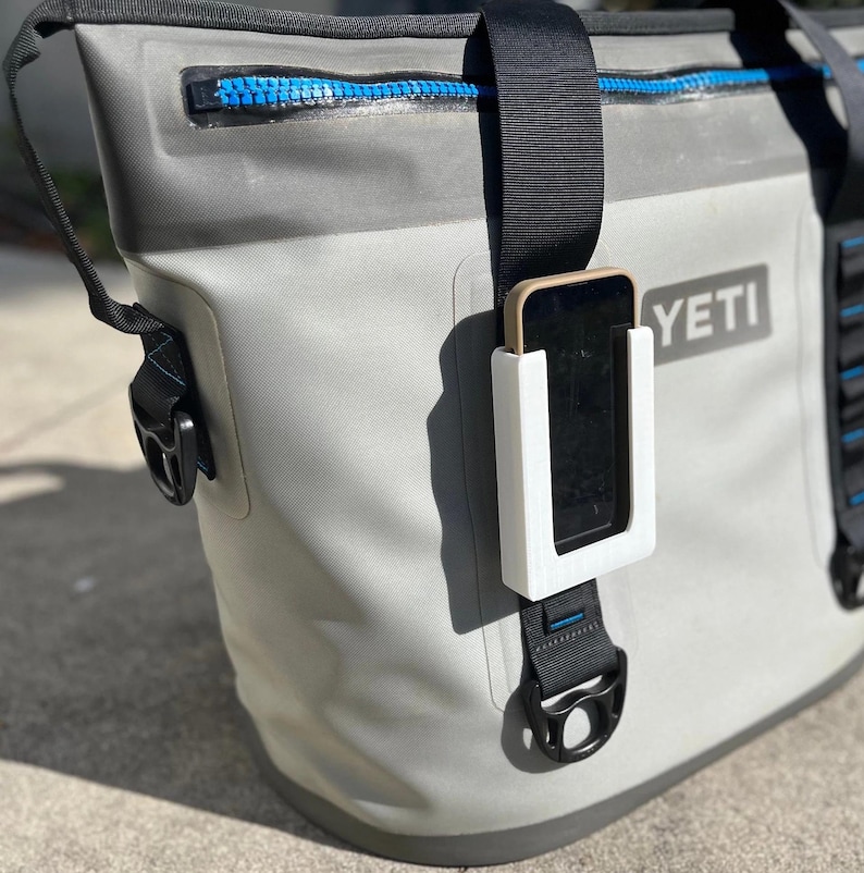 Puede incluir: Una bolsa de enfriador Yeti gris con una correa negra y un soporte de tel&eacute;fono blanco unido a la correa. El soporte de tel&eacute;fono tiene un recorte para un tel&eacute;fono y un tel&eacute;fono negro es visible dentro del soporte. La bolsa de enfriador tiene una cremallera azul y la palabra "YETI" est&aacute; impresa en el lateral.