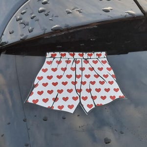 Könnte beinhalten: Ein weißer Aufkleber in Form einer Boxershorts mit einem wiederkehrenden Muster aus kleinen roten Herzen, der auf der dunkel getönten Heckscheibe eines Fahrzeugs angebracht ist. Der Aufkleber hat schwarze Umrisse für die Form.