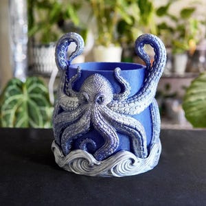 May include: A cylindrical blue planter featuring a raised, silver-toned octopus relief wrapped around the exterior. The octopus has detailed suckers on its tentacles, which extend upward, and the base is decorated with sculpted, swirling white waves.
