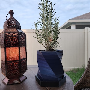 May include: A dark blue, geometric planter with a rosemary plant growing inside. The planter has a hexagonal base and a twisted, cylindrical design. The planter is sitting on a wooden table next to a decorative lantern.