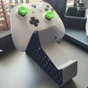 May include: White Xbox controller with green thumbsticks resting on a white and navy blue controller stand. The stand has a honeycomb pattern on the front and a curved design to hold the controller securely.