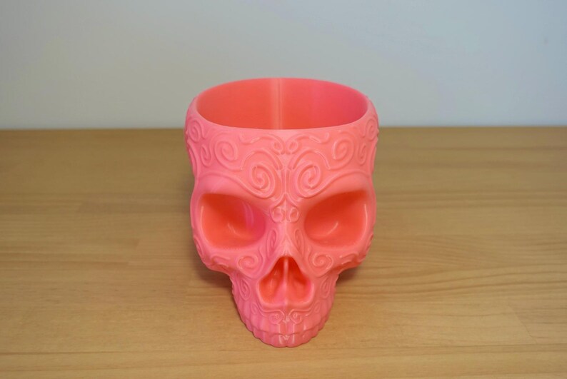 May include: A pink skull-shaped planter with intricate, swirling patterns. The decorative pot has a wide opening and is made of a solid, smooth material. The planter is designed to hold small plants or succulents.