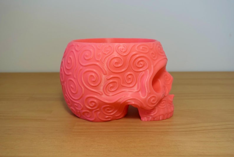 May include: A coral-colored skull-shaped planter with a detailed, swirling pattern. The decorative planter is made of a solid material and sits on a wooden surface. The skull has an open top for planting.