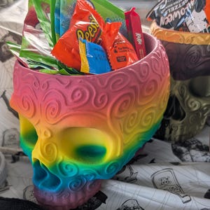 May include: A rainbow-colored skull-shaped container filled with various wrapped candies. The container has a detailed, swirling design and is painted in a gradient of colors, including pink, yellow, green, and blue. The candies include Reese's, Kit Kat, and other assorted treats.