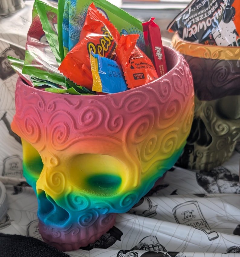 May include: A rainbow-colored skull-shaped container filled with various wrapped candies. The container has a detailed, swirling design and is painted in a gradient of colors, including pink, yellow, green, and blue. The candies include Reese's, Kit Kat, and other assorted treats.