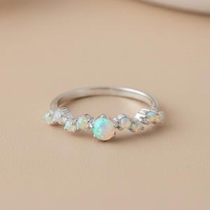 May include: A silver-toned ring featuring a central round opal flanked by three smaller opals on each side. The stones are set in a prong setting along the top half of the thin, polished metal band, showcasing iridescent flashes of color.