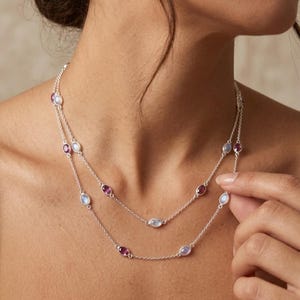 May include: A double-strand silver necklace featuring alternating oval-shaped moonstones and deep pink gemstones. The delicate chain links the stones in a spaced pattern, creating a layered look around the neck with a polished metallic finish.