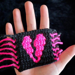 Puede incluir: Un parche de coraz&oacute;n de ganchillo negro y rosa con flecos en los bordes.