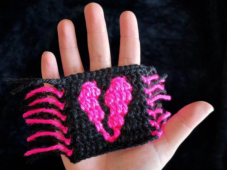 Puede incluir: Un parche de coraz&oacute;n de ganchillo negro y rosa con flecos en los bordes.