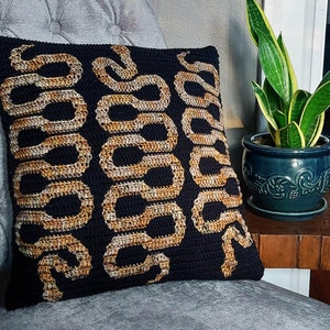 May include: A black and gold crocheted pillow with a snake pattern. The pillow is sitting on a gray couch.