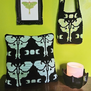 May include: A black crochet cushion and tote bag featuring a moth design in light green. A framed moth print hangs on the wall above the cushion. A black candle holder with a pink candle sits on a wooden surface.