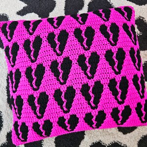 Puede incluir: Una almohada de crochet rosa con un patr&oacute;n negro de corazones repetidos.