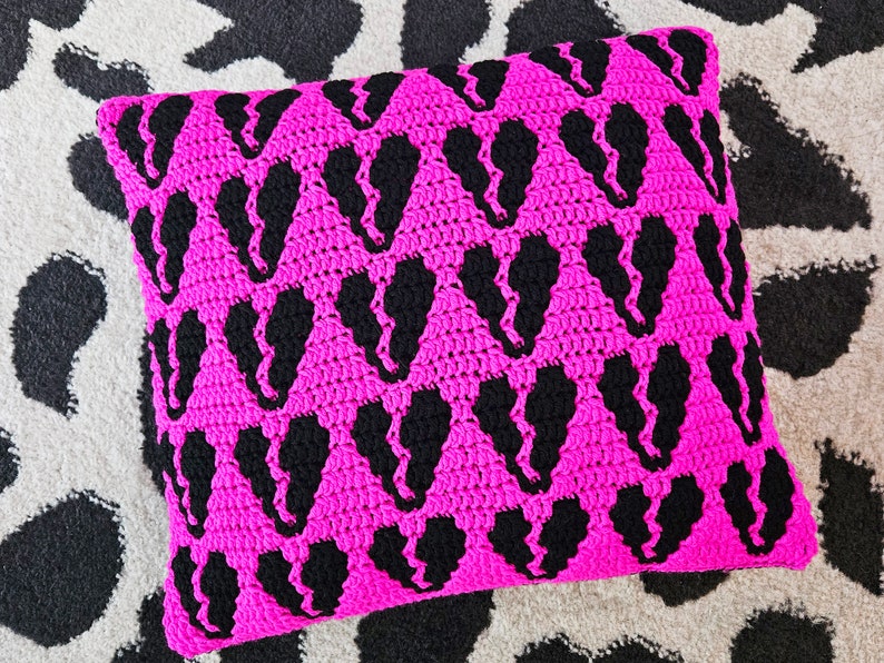 Puede incluir: Una almohada de crochet rosa con un patr&oacute;n negro de corazones repetidos.