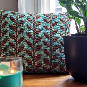 Puede incluir: Una almohada de crochet azul turquesa y marrón con un patrón de hojas se encuentra sobre una superficie de madera junto a una vela azul turquesa y una maceta negra con una planta verde.