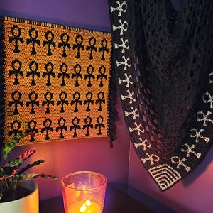May include: A black and white crocheted shawl with an ankh symbol pattern. The shawl is hanging on a purple wall next to a wall hanging with a repeating pattern of ankh symbols in black and yellow.