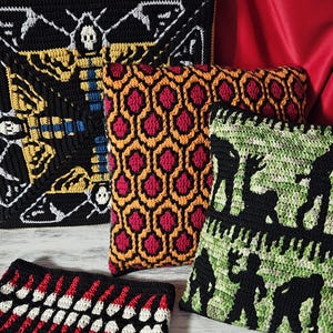 May include: A collection of crocheted pillows with various designs. One pillow features a black and yellow moth design with a skull. Another has a red and orange honeycomb pattern. A third pillow displays black silhouettes on a green background. The pillows are on a white surface.