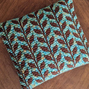 May include: A brown and blue crocheted cushion with a leaf pattern. The cushion is on a wooden surface.