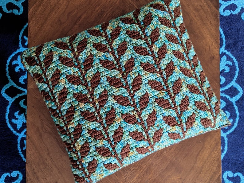 May include: A brown and blue crocheted cushion with a leaf pattern. The cushion is on a wooden surface.