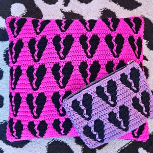 Puede incluir: Una almohada de crochet rosa con un patr&oacute;n de coraz&oacute;n negro y una bolsa de crochet morada a juego con un patr&oacute;n de coraz&oacute;n negro.