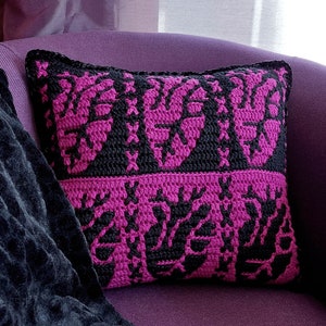 May include: A pink and black crocheted throw pillow with a geometric pattern. The pillow is sitting on a purple couch.