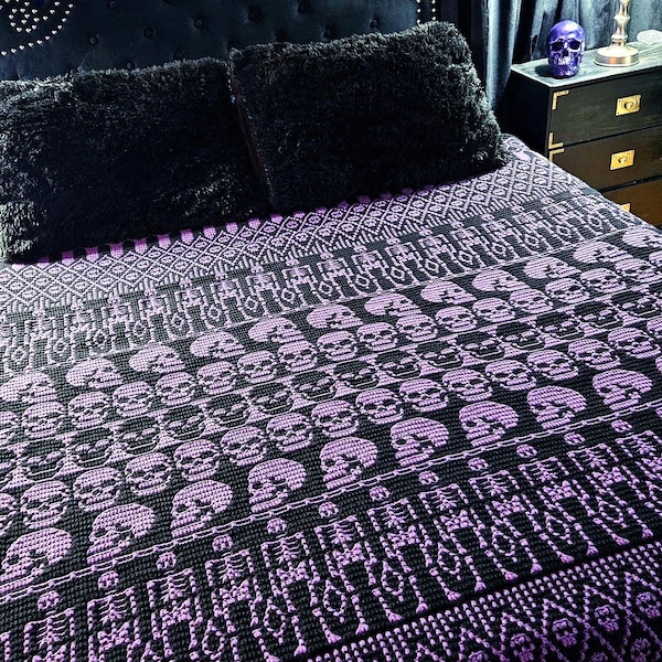 May include: A black and purple knit blanket featuring a repeating pattern of skulls and skeletons. The blanket is draped over a bed with two black faux fur pillows. The design includes geometric borders and rows of skulls in a dark color scheme.
