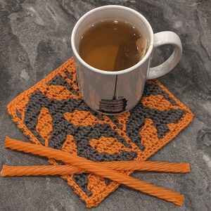 May include: A close-up of a crocheted coaster with an orange and black pattern. A white mug with tea and a tea bag is sitting on the coaster. Two orange liquorice twists are on top of the coaster.