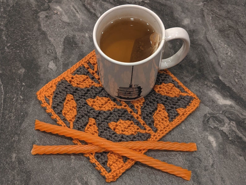 May include: A close-up of a crocheted coaster with an orange and black pattern. A white mug with tea and a tea bag is sitting on the coaster. Two orange liquorice twists are on top of the coaster.