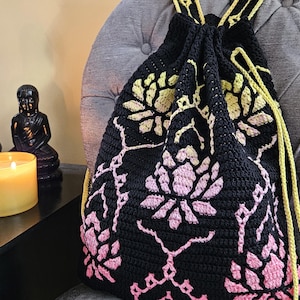May include: Black crochet drawstring backpack with a floral design in pink and yellow. The bag has yellow drawstrings and straps. The bag is sitting on a gray upholstered chair.