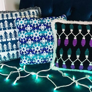 Puede incluir: Tres fundas de cojín de crochet con temas navideños de invierno. La primera funda de cojín presenta un patrón blanco y azul de pinos estilizados. La segunda funda de cojín tiene un patrón geométrico azul y blanco. La tercera funda de cojín tiene un fondo negro con un patrón blanco y verde de bombillas estilizadas con acentos morados.