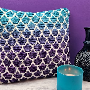 Peut inclure: Un coussin crocheté avec un motif d'écailles de sirène dans des tons de violet, bleu et blanc. Le coussin est posé sur une surface blanche à côté d'une bougie bleue et d'un vase noir.