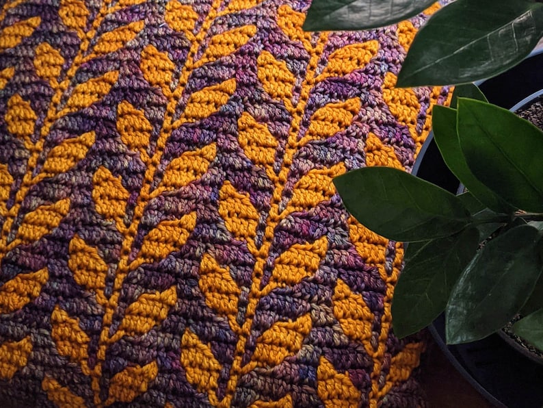 May include: A close-up of a crocheted throw blanket featuring a repeating pattern of yellow and purple leaves. The blanket is crafted with a textured yarn and has a soft, cosy feel.