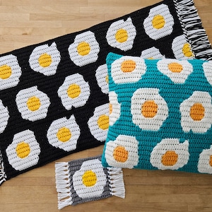 May include: Three crocheted throw cushions with a pattern of white eggs with yellow yolks on a black, teal, and grey background. The cushions have fringe around the edges.