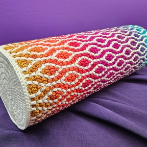Peut inclure: Un coussin cylindrique en crochet coloré avec un fond blanc et un motif géométrique. Le coussin est fait avec un dégradé de couleurs, commençant par l'orange et passant au violet.