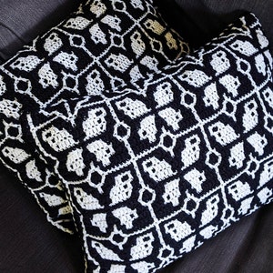 Peut inclure: Deux coussins de décoration en crochet noir et blanc avec un motif géométrique de fantômes. Les coussins sont sur un canapé gris foncé.