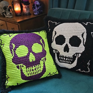Puede incluir: Dos cojines cuadrados de ganchillo con borde negro difuso. Uno presenta una calavera morada sobre un fondo verde lima. El segundo muestra una calavera blanca sobre un fondo negro. Ambos cojines tienen un diseño de calavera detallado y cosido.