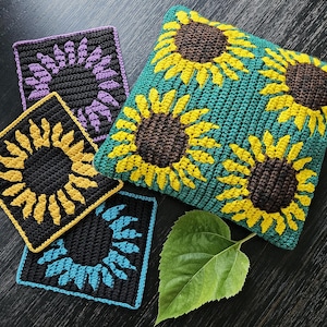May include: Four crocheted sunflower squares in purple, yellow, blue, and green. The squares are arranged on a dark wooden surface with a green leaf.