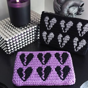 Puede incluir: Dos estuches de cremallera de ganchillo con un dise&ntilde;o de coraz&oacute;n roto en blanco y negro. Un estuche es morado y el otro es negro.