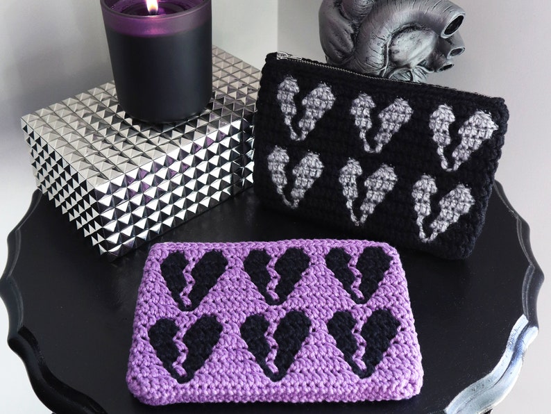 Puede incluir: Dos estuches de cremallera de ganchillo con un dise&ntilde;o de coraz&oacute;n roto en blanco y negro. Un estuche es morado y el otro es negro.