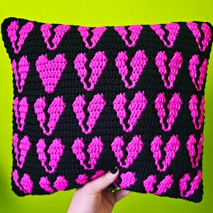 Puede incluir: Una almohada de crochet negra con un patr&oacute;n repetido de corazones rosas.