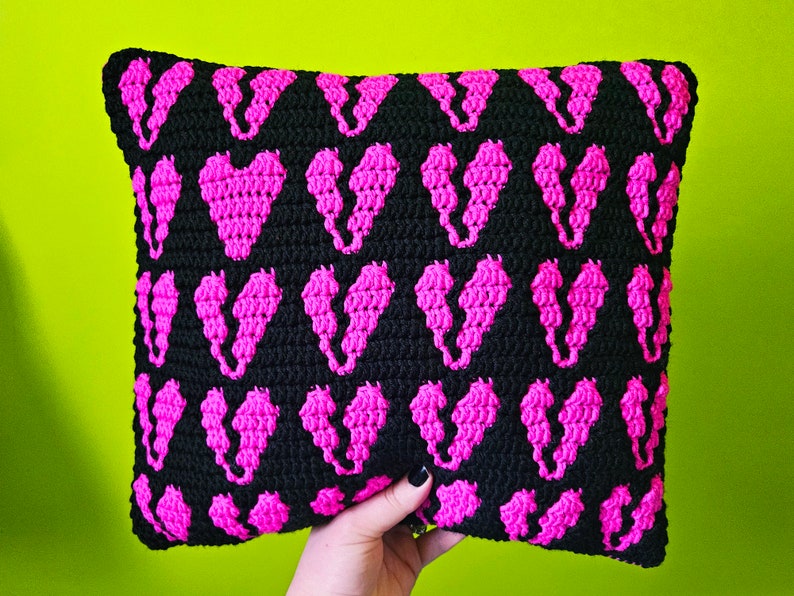 Puede incluir: Una almohada de crochet negra con un patr&oacute;n repetido de corazones rosas.