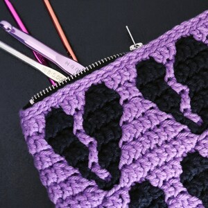 Puede incluir: Bolsa de cremallera de crochet morado con detalles de crochet negro. La bolsa est&aacute; abierta y tiene ganchos de crochet en su interior.