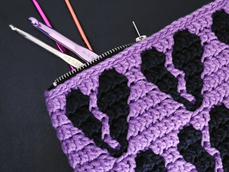 Puede incluir: Bolsa de cremallera de crochet morado con detalles de crochet negro. La bolsa est&aacute; abierta y tiene ganchos de crochet en su interior.