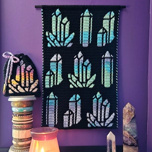 May include: A crocheted wall hanging with a black background and colorful crystal designs. The hanging is displayed on a purple wall with a crocheted pouch and crystal points on a table below.