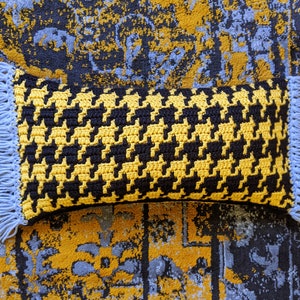 May include: A rectangular, handmade crochet pillow with a black and yellow houndstooth pattern. The pillow has light blue fringe on each end. The pillow is placed on a patterned rug with blue, yellow, and black colors.